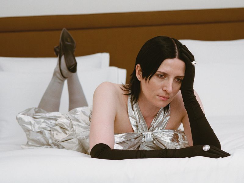 Adult woman lies on white bed in silver dress and black gloves, looking toward camera in a portrait fashion photograph.