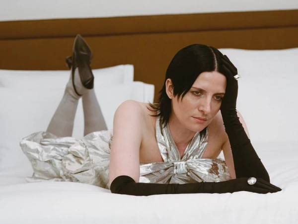 Adult woman lies on white bed in silver dress and black gloves, looking toward camera in a portrait fashion photograph.