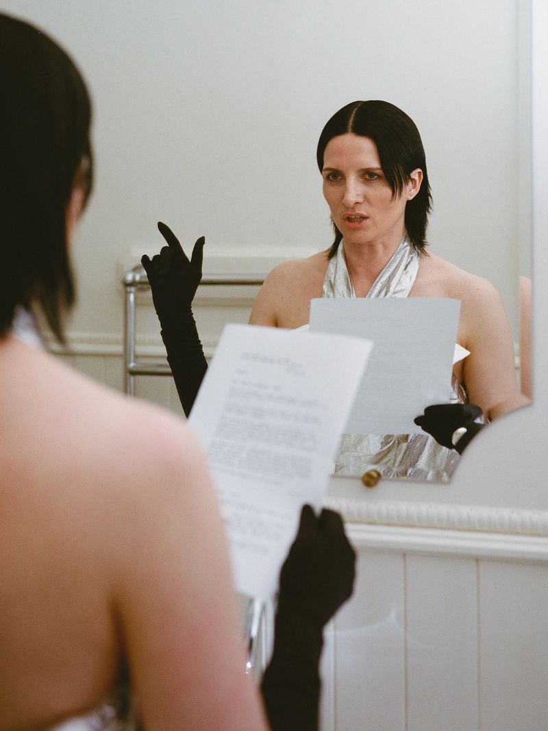 Woman in a silver top and black gloves is seen in a mirror, holding and reading papers in a white tiled bathroom setting.