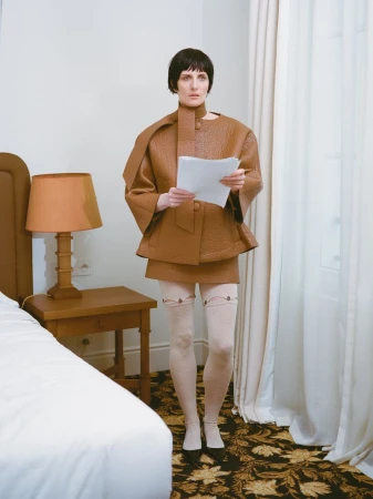 Woman in brown jacket and skirt holds papers indoors near window, bed, and lamp. Neutral indoor fashion portrait.