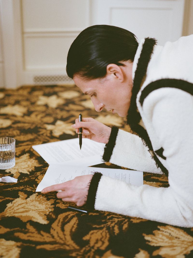 Woman in a white robe lying on a patterned carpet, focused on papers in front of her, holding a pen, with a glass of water nearby.