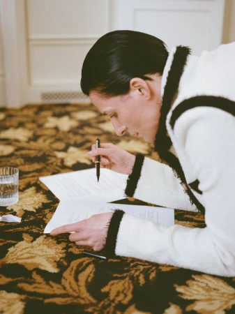 Woman in a white robe lying on a patterned carpet, focused on papers in front of her, holding a pen, with a glass of water nearby.
