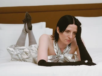 Model Kim Peers lies on a white bed wearing a metallic silver dress, long black opera gloves, and black slingback heels.