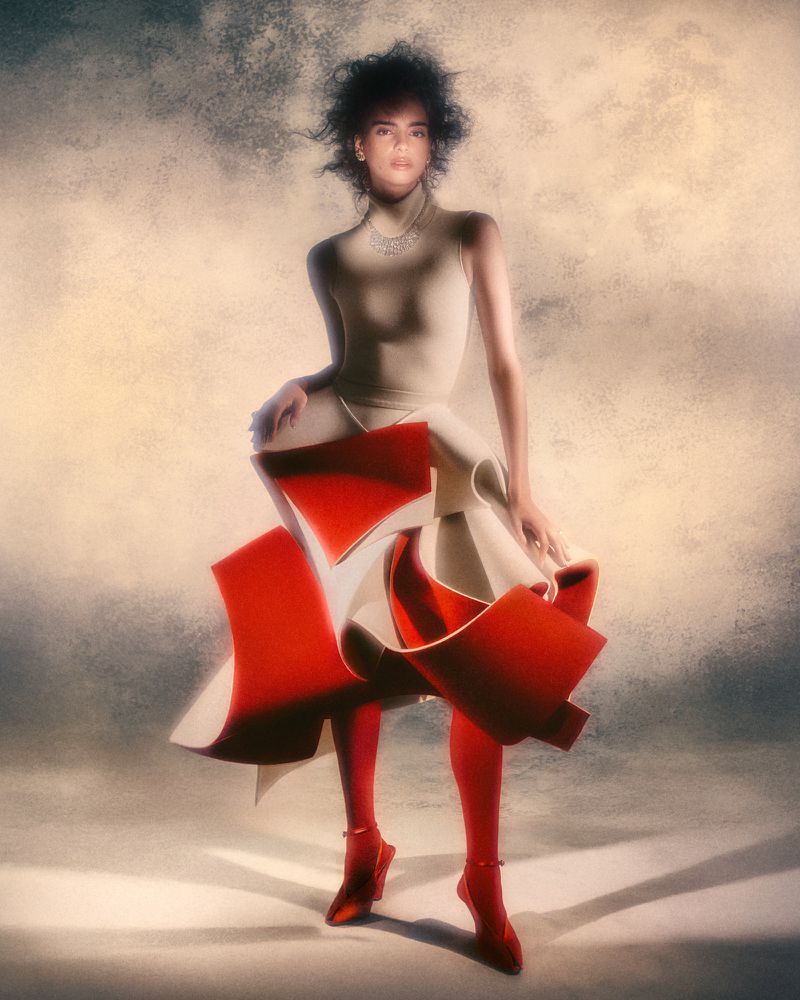 Elegant woman in a cream-colored sleeveless bodysuit, architectural layered skirt, red tights, and heels posing against a painted background.