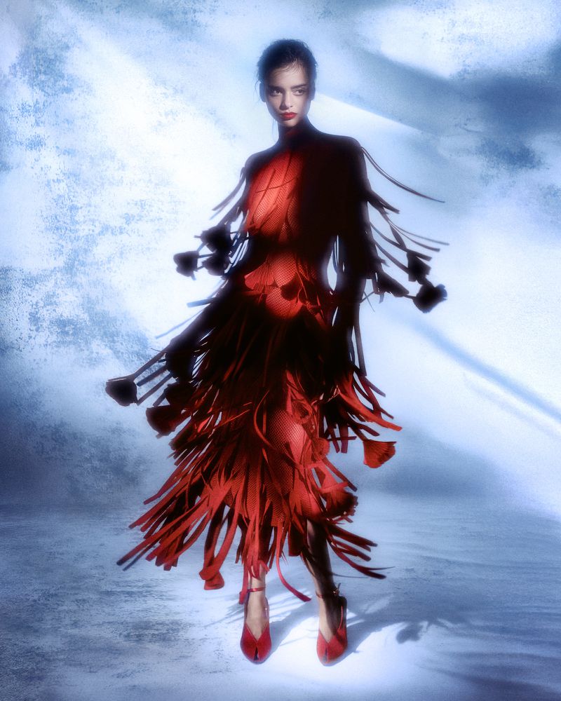 Fashion model in a red dress with fringed details poses in a photo studio with a blurred blue and white background and dramatic lighting.