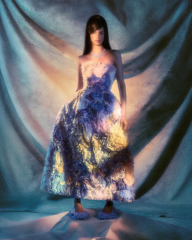 A woman with dark hair is posing in a fluffy, strapless gown with matching slippers in a studio with textured fabric backdrop.
