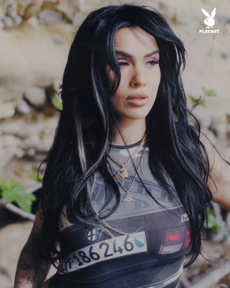Close-up shot of an adult woman with dark hair and a printed tank top, outdoors with Playboy logo in the upper right corner.