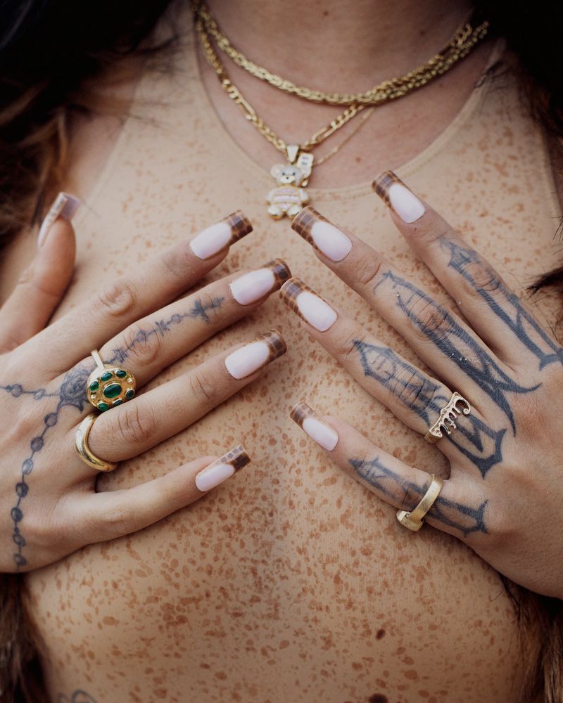 Close-up studio shot of a person's chest and tattooed hands adorned with rings and pink nails, accented by layered gold necklaces.
