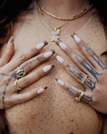 Close-up studio shot of a person's chest and tattooed hands adorned with rings and pink nails, accented by layered gold necklaces.