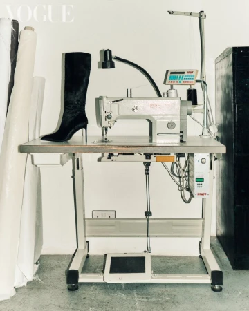 Fashion shot of a black leather stiletto boot on a sewing machine table in a studio setting with white fabric rolls in the background.