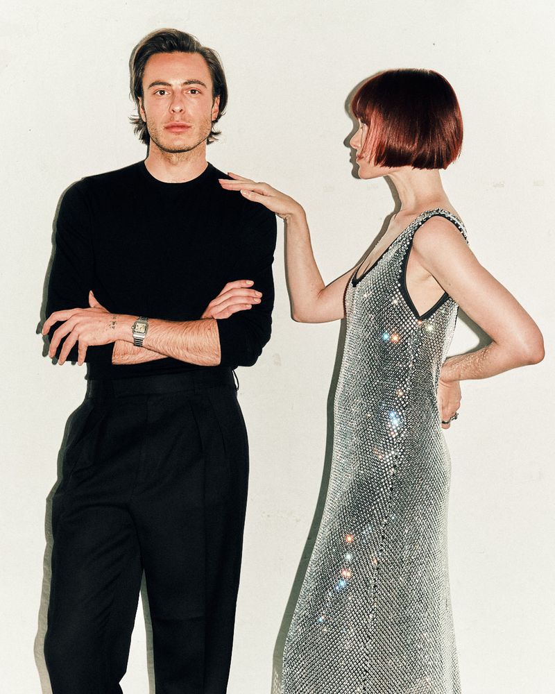A man and a woman in a studio setting. The man wears black attire, while the woman is in a silver dress, posing with her hand on his shoulder.