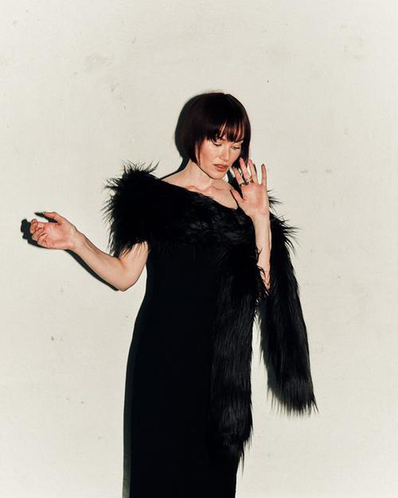 Elegant woman with bobbed hair in a black dress and boa stands against a creamy white wall in a studio setting.