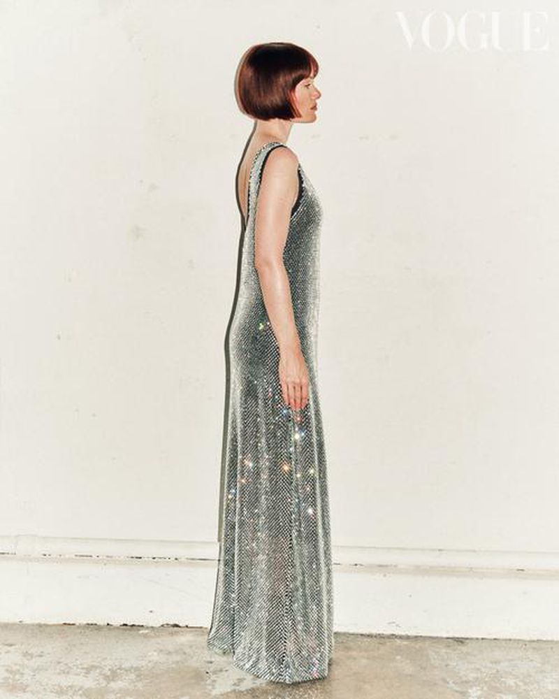 A woman in a silver sequin gown stands in profile next to a white wall, fashion editorial with a bob haircut, full length shot.