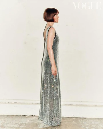 A woman in a silver sequin gown stands in profile next to a white wall, fashion editorial with a bob haircut, full length shot.