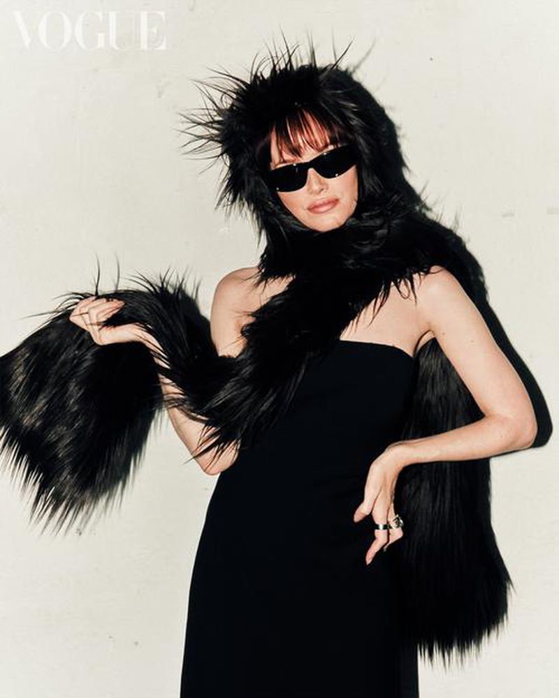 Model wearing black sunglasses, strapless black dress, and a long shaggy black faux fur stole against a pale wall.