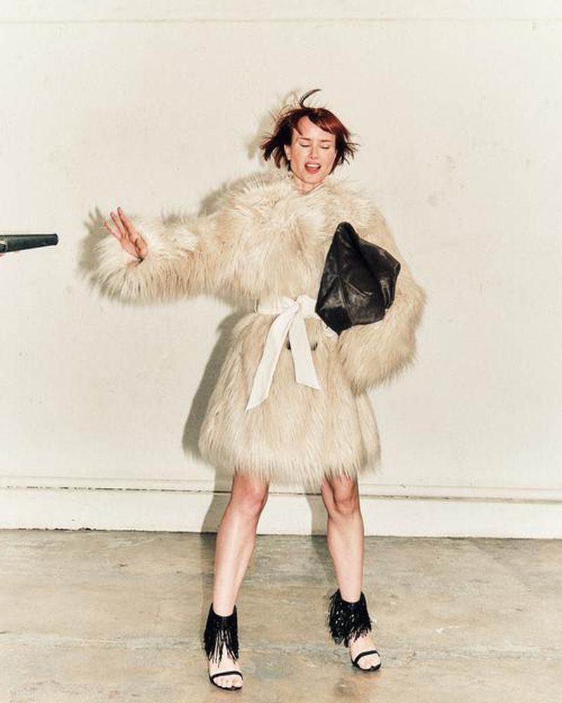 Woman in a fur coat striking a pose with a bag in a light studio setting, wearing high heels with fringe, against a white background.
