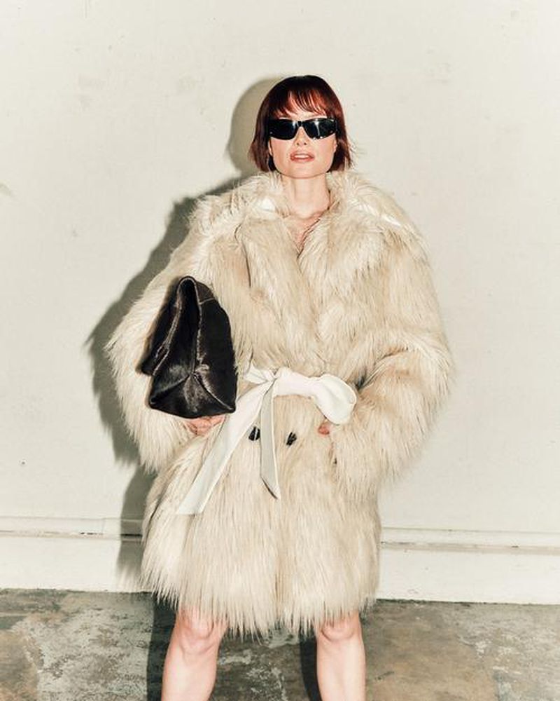 Woman in a shaggy faux fur coat, sunglasses, and holding a handbag poses against a concrete wall, fashion portrait indoors.