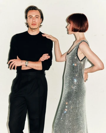 Man in black crewneck sweater with arms crossed standing next to a woman in a sparkling silver dress