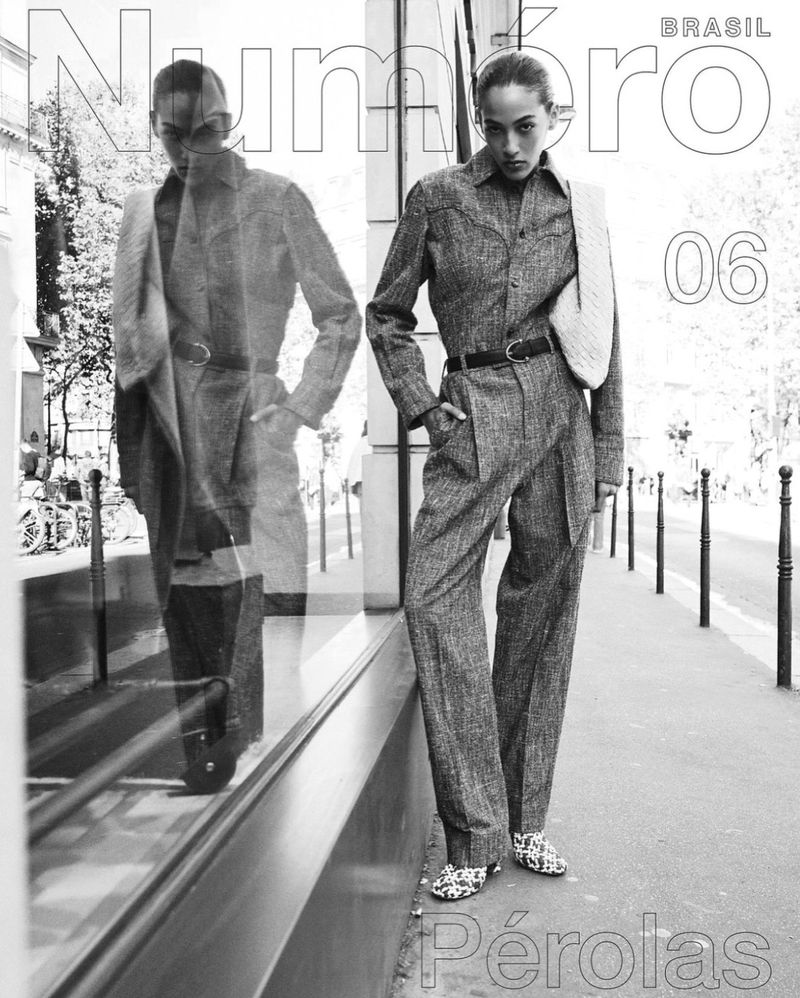 Fashion editorial shot. An adult female model in a gray jumpsuit stands on a city sidewalk next to a building with reflective windows.