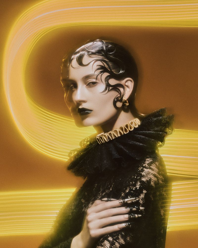 A woman with styled dark hair and dramatic makeup poses in a lace top with a ruffled collar against a blurred yellow background.