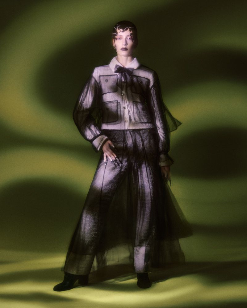 A person in a sheer black and white outfit stands in front of a blurred green backdrop, bathed in soft light. Fashion, portrait.