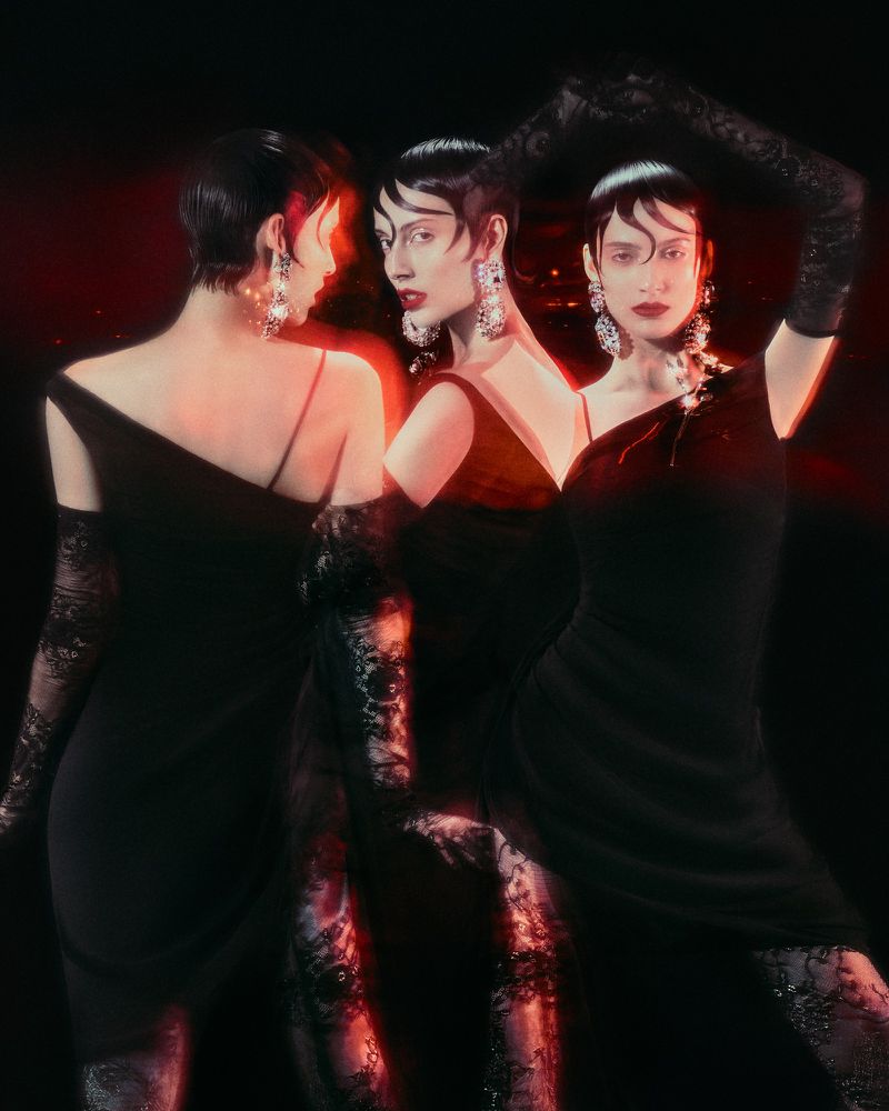 Studio shot of a woman with short hair wearing a black dress, gloves and dangling earrings against a dark background with red light.