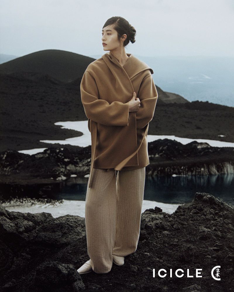 Fashion shot of a woman in a tan coat and pants, standing on a rocky landscape with a snowy background, looking away pensively.
