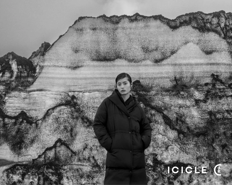 Monochrome portrait of an Asian woman in a puffer jacket standing in front of a stratified rock formation under a cloudy sky.