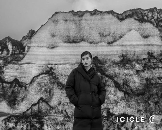 Monochrome portrait of an Asian woman in a puffer jacket standing in front of a stratified rock formation under a cloudy sky.