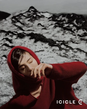 A woman in a red hooded garment poses outdoors against a stark landscape of snow-dusted rocks, creating a striking contrast in color and texture.