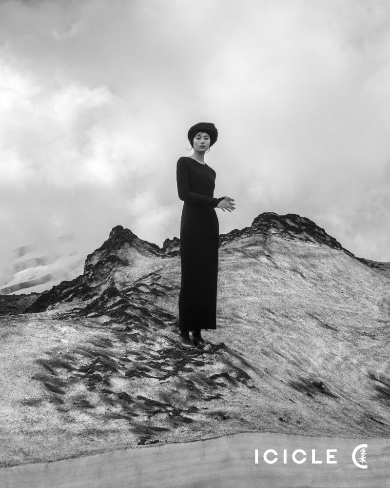 Monochrome fashion shot of a woman in a long black dress and hat posing on a rocky landscape under a cloudy sky, stylized by ICICLE.