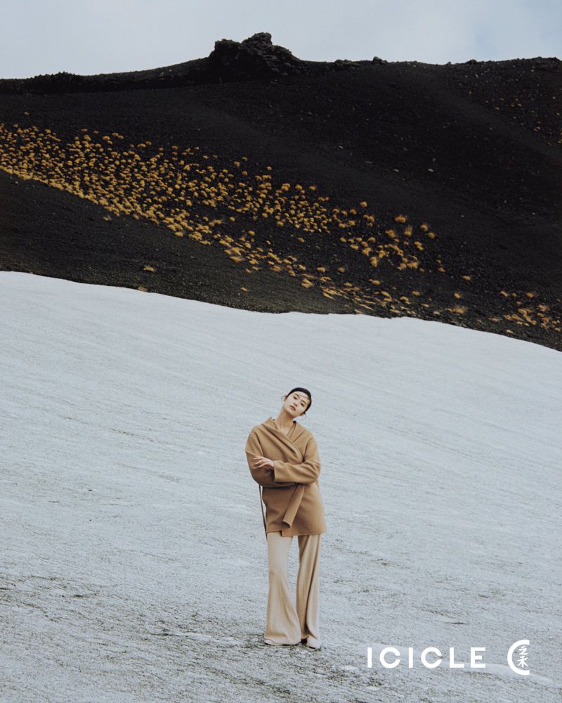 A full shot of a female model posing in a tan coat and pants on an ice field in front of a black, grassy mountain under an overcast sky.