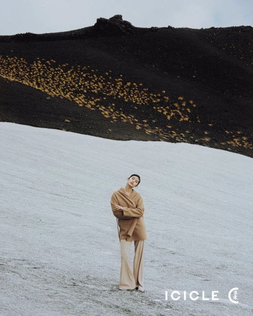 A full shot of a female model posing in a tan coat and pants on an ice field in front of a black, grassy mountain under an overcast sky.