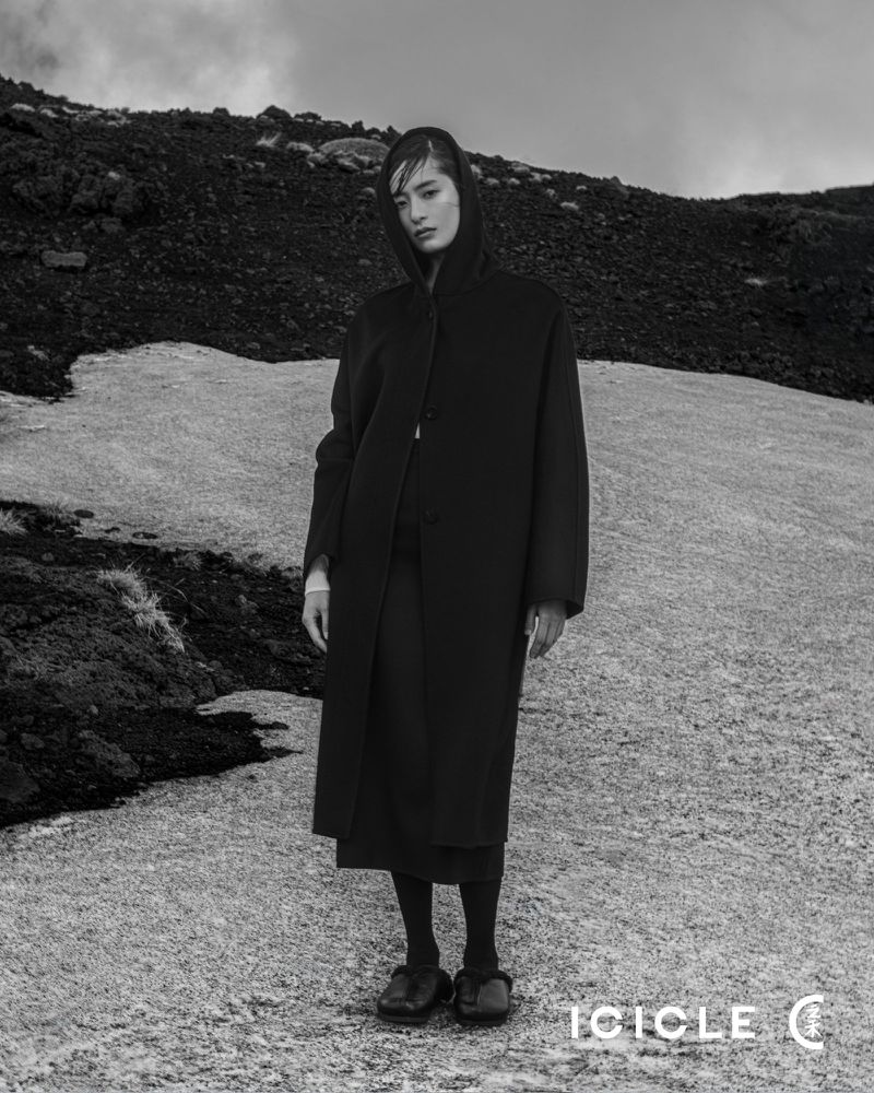 Monochrome fashion portrait of a woman in a hooded coat posing against a rocky hillside with snow accents on an overcast day.