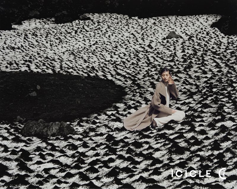 Woman wearing a long coat sits cross-legged in a field with a patterned ground and dark vegetation in a black-and-white fashion shot.