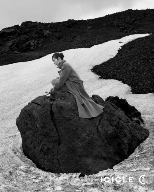 A woman in a coat sits on a rock in a snowy, mountainous landscape. The black and white image evokes a sense of solitude and natural beauty.