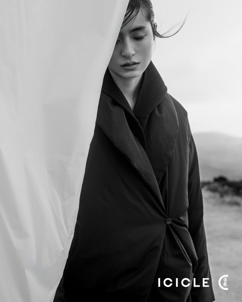 Elegant portrait of an Asian female model in a black coat with white fabric accent, set against a muted outdoor background in soft light.