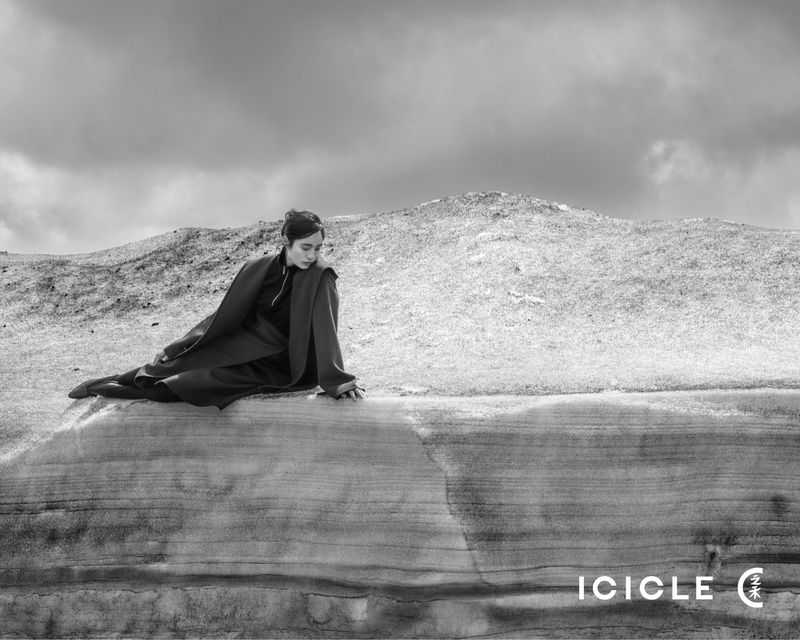 A woman poses in a long coat on a ridge in a monochrome outdoor fashion shot against a background of rolling hills and cloudy sky.