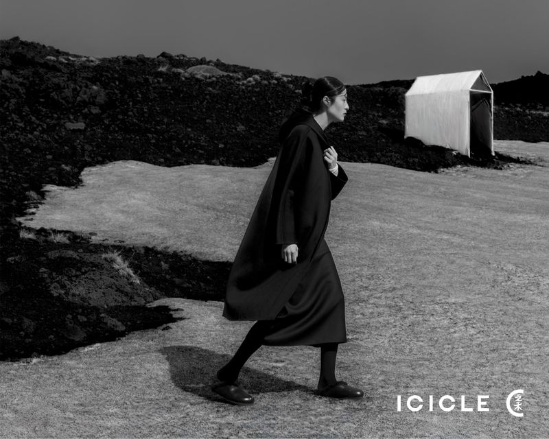 Monochrome outdoor fashion shot: An adult woman in a dark coat walks across a rugged landscape toward a tent-like structure under an overcast sky.