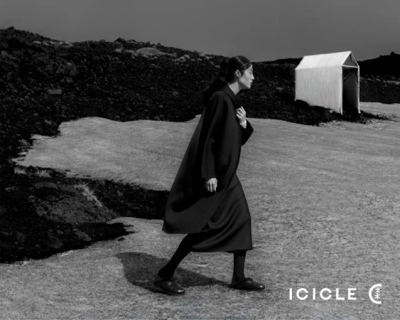 Monochrome outdoor fashion shot: An adult woman in a dark coat walks across a rugged landscape toward a tent-like structure under an overcast sky.