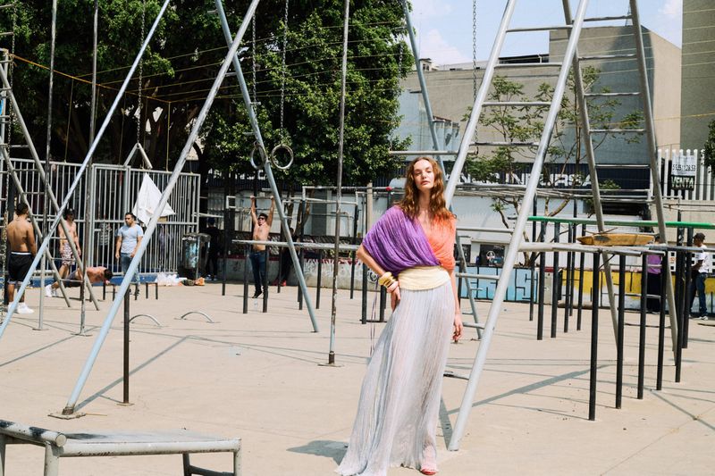 Fashion shot of woman in colorful draped top and flowing skirt posing by metal jungle gym structure in urban park setting with other people.