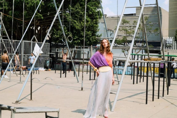Fashion shot of woman in colorful draped top and flowing skirt posing by metal jungle gym structure in urban park setting with other people.