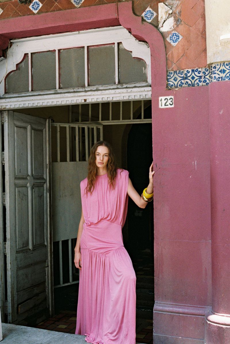 A woman in a pink dress poses by a building entrance with decorative tiles. She wears bracelets and looks at the camera.