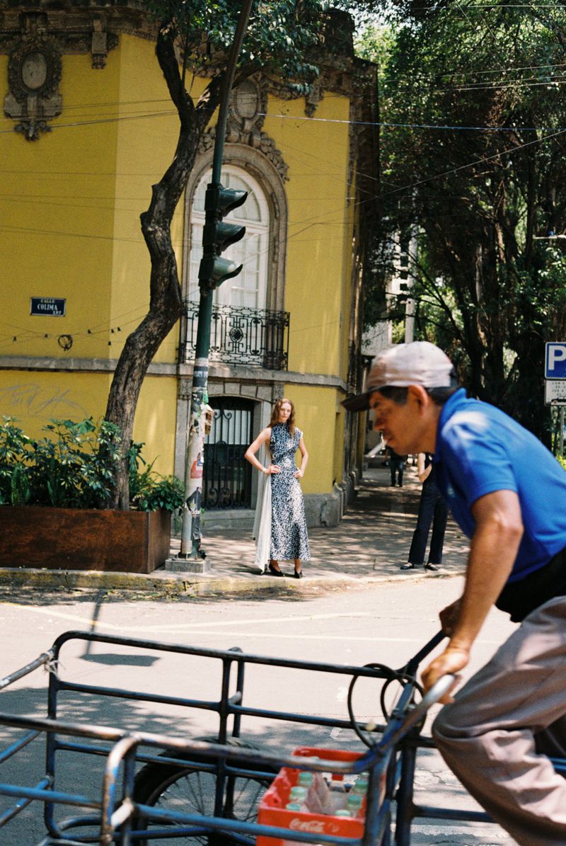 Model in patterned dress poses on city street near pedestrian with cart, set against yellow building and green trees in daylight.