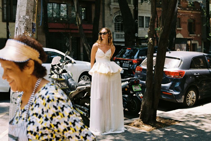 Woman in a dress standing on a city sidewalk with other pedestrians and parked cars visible in the background on a sunny day.