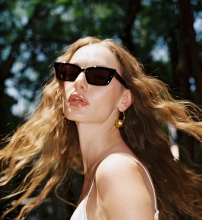 Danielle van Randen wearing black sunglasses and a gold sphere earring with long wavy hair blowing in the sunlight.
