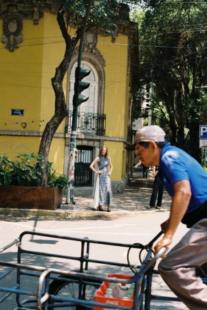 Model posing in a patterned gown by a yellow building as a man rides past on a cargo tricycle.