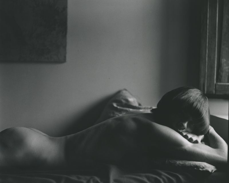 Black and white photograph of a woman with a dark bob haircut lying prone on a bed near a window with soft directional light.