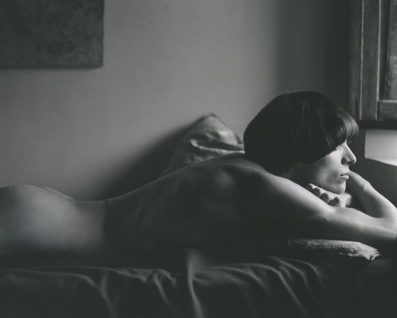 Monochrome portrait of a woman with a bowl cut lying on her stomach with her head propped on her hands, looking out a window.
