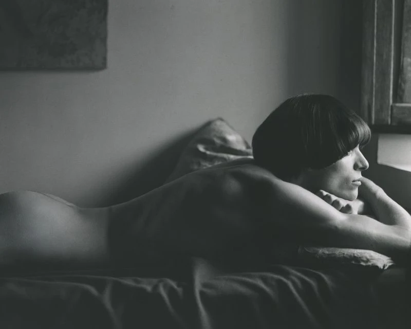 Monochrome portrait of a woman with a bowl cut lying on her stomach with her head propped on her hands, looking out a window.